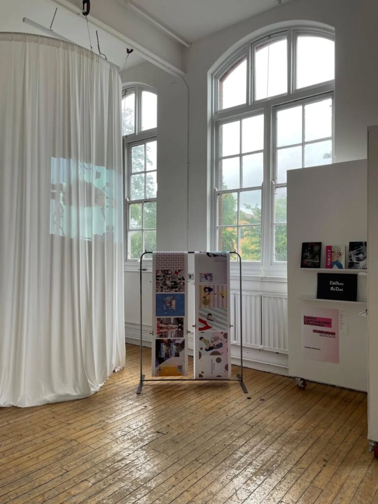 A bright room with large windows, wooden floors, a white curtain with a projected image, a standing display board with colorful images, and a white shelf holding books and papers.