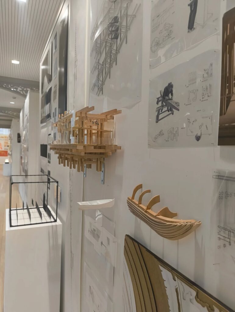 Architectural models and technical drawings are displayed on white walls, featuring wooden and metal structures, diagrams, and sketches. The exhibit showcases detailed construction and design concepts in a well-lit gallery space.