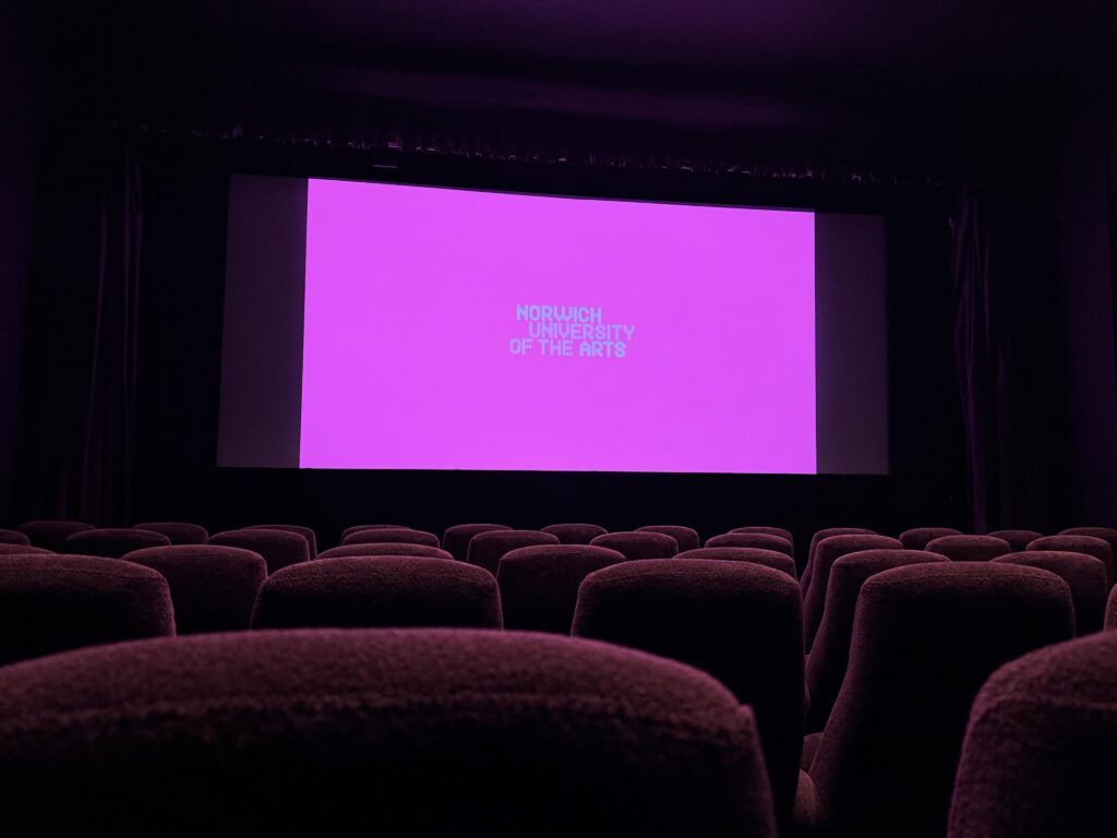 A dark movie theater with rows of empty seats facing a large screen displaying a bright magenta background and the words "Norwich University of the Arts" in the center.