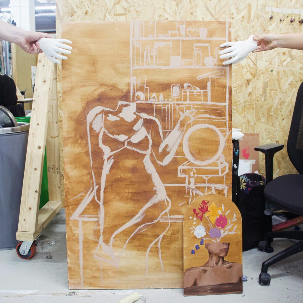 Two people hold a large unfinished painting of a seated, faceless figure in lingerie, with a bookshelf background sketched in white. A smaller painting of a torso with a bouquet for a head leans against it.