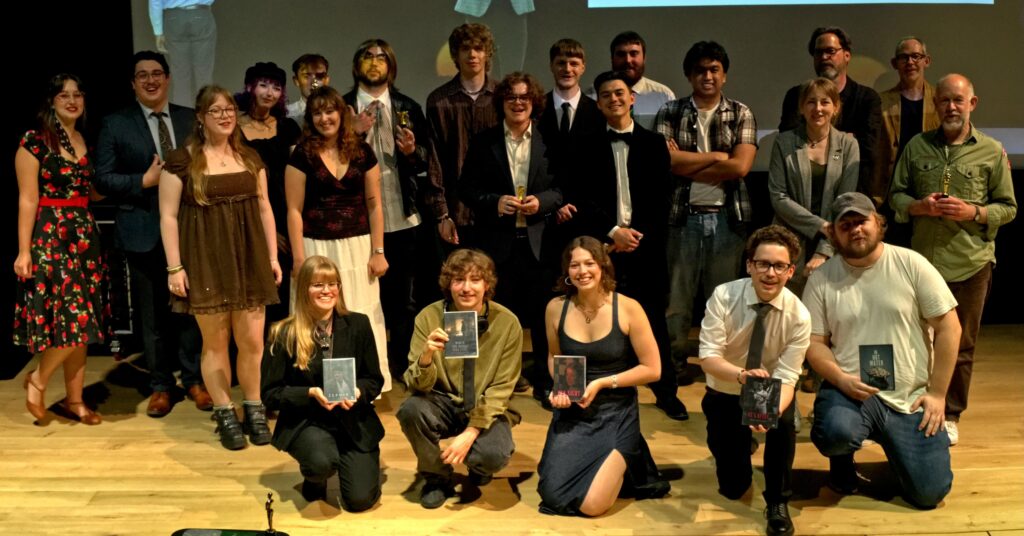 A group of people posed on a wooden stage, some standing and others kneeling in front, with a few holding glass awards and all smiling toward the camera. There's a presentation screen in the background.
