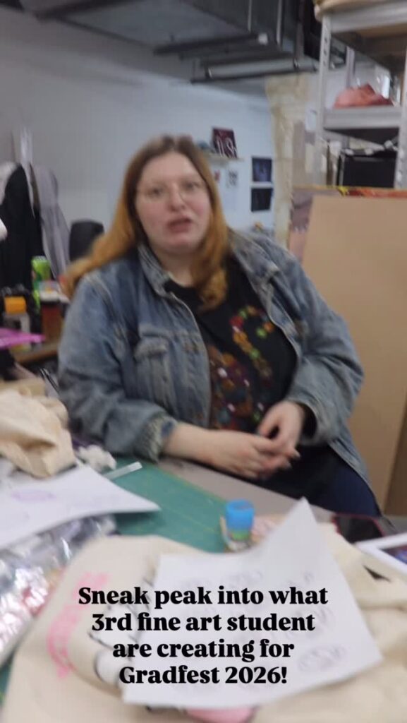 A person with long hair and glasses, wearing a denim jacket, sits at a cluttered art desk. Art supplies and papers are scattered in the foreground. Text on the image reads: "Sneak peak into what 3rd fine art student are creating for Gradfest 2026!.