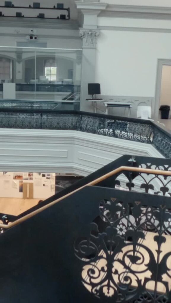Ornate black metal railing lines a staircase and balcony inside a bright building with white walls, large windows, and display boards on the lower floor. Modern furniture sits near the glass-walled upper area.