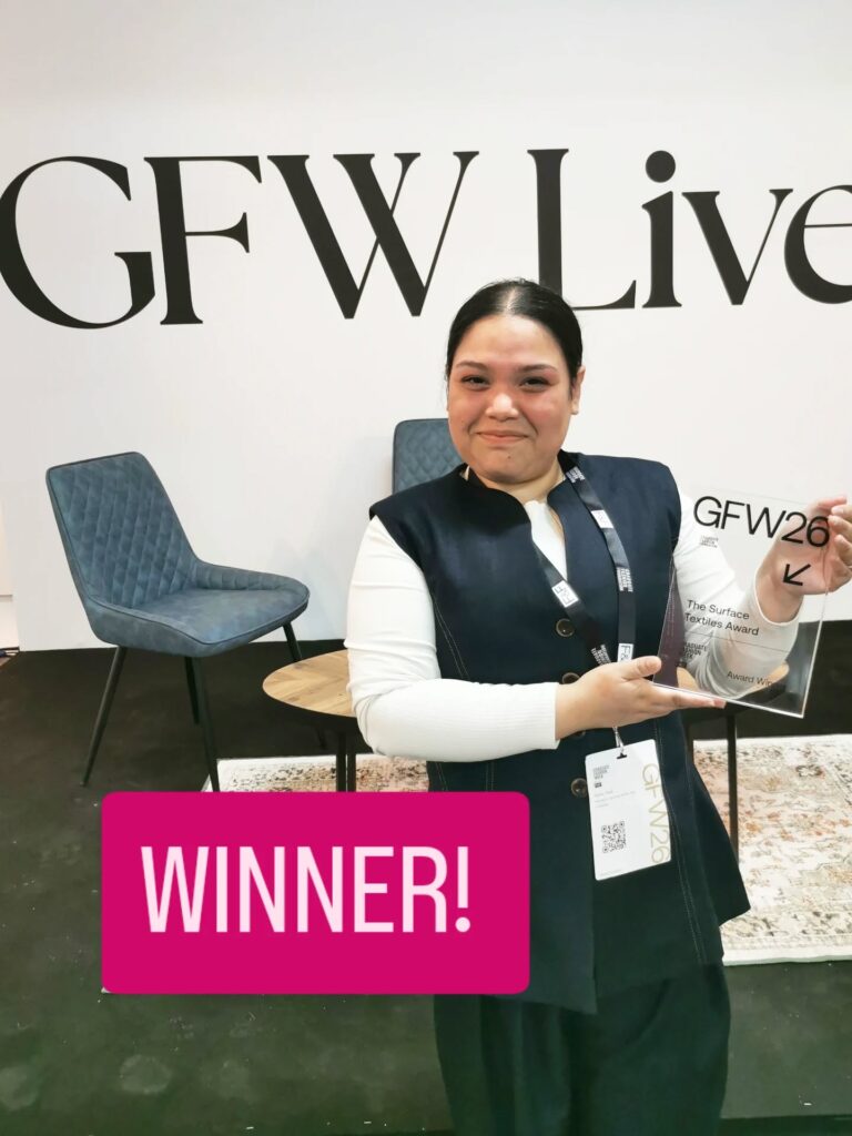 A smiling woman stands indoors, holding a clear award plaque labeled "GFW26." Behind her are modern chairs and a table, with a white wall displaying the text "GFW Live." The image features a bold "WINNER!" graphic.