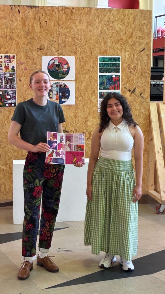Two young women stand indoors by a display board with colorful art. One holds a collage; she wears floral pants and a dark shirt. The other wears a white top and green checkered skirt. Artworks are mounted on the wooden board behind them.