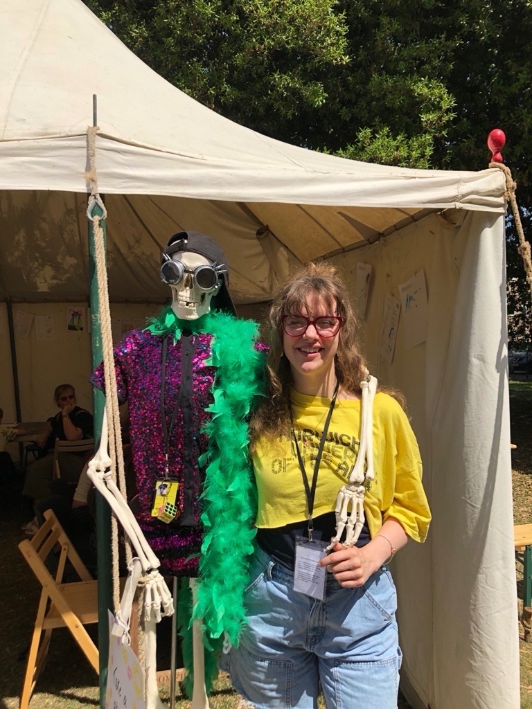 A smiling person wearing a yellow shirt and name badge stands next to a skeleton dressed in a hat, sunglasses, a purple sequin top, and a green feather boa, both posing in front of a white tent in a sunny outdoor setting.