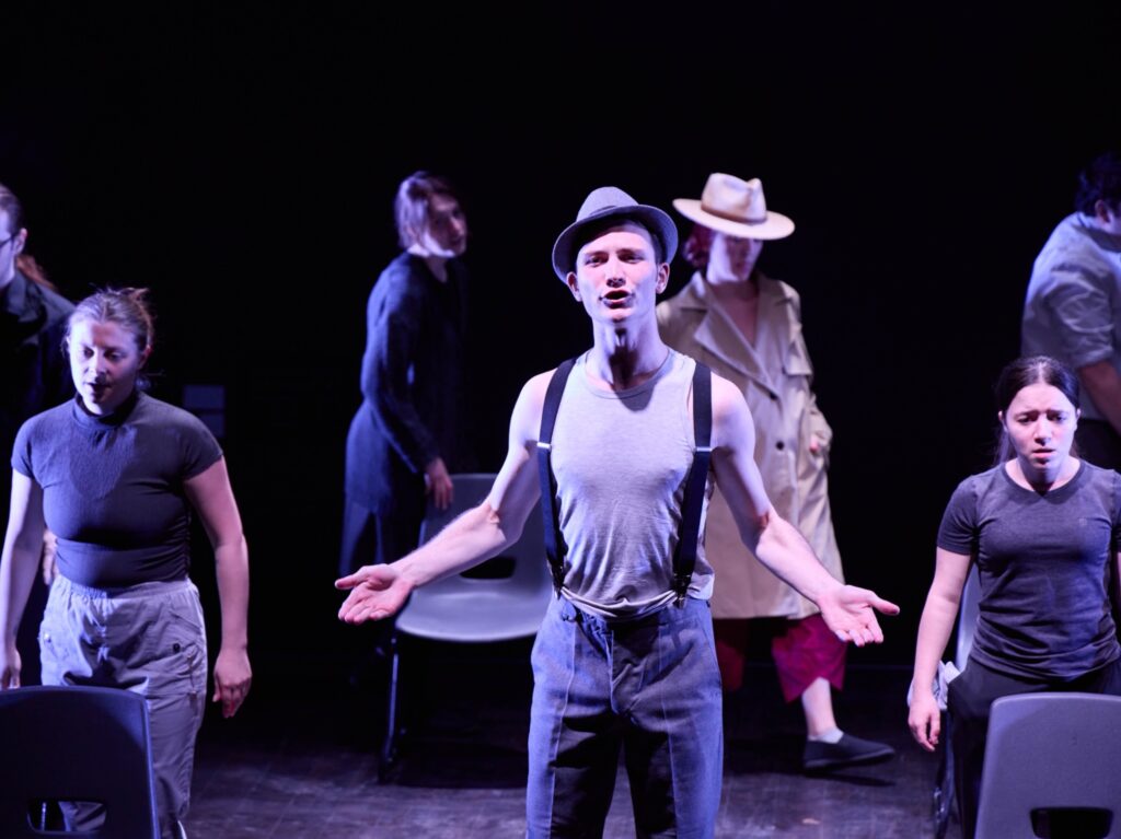 A group of actors perform on stage under dramatic lighting. The central figure wears suspenders, a gray hat, and a gray shirt, with arms open, while others stand around, some in shadow and one in a trench coat and fedora.