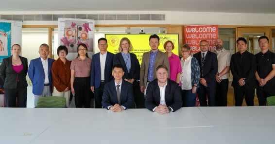 A group of fifteen people, including men and women in business attire, pose together in a meeting room. Two men are seated at a white table while the others stand behind them. Banners and posters are visible in the background.