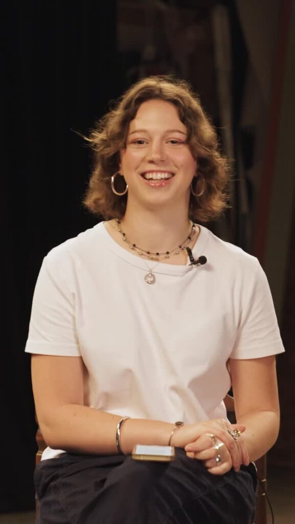 A young woman with short wavy hair, wearing a white t-shirt and jewelry, sits smiling with a microphone clipped to her shirt against a dark background.