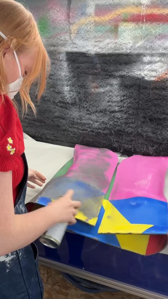 A girl wearing a face mask and red sweater spray paints colorful designs, including a yellow star, on pieces of paper laid out on a table.