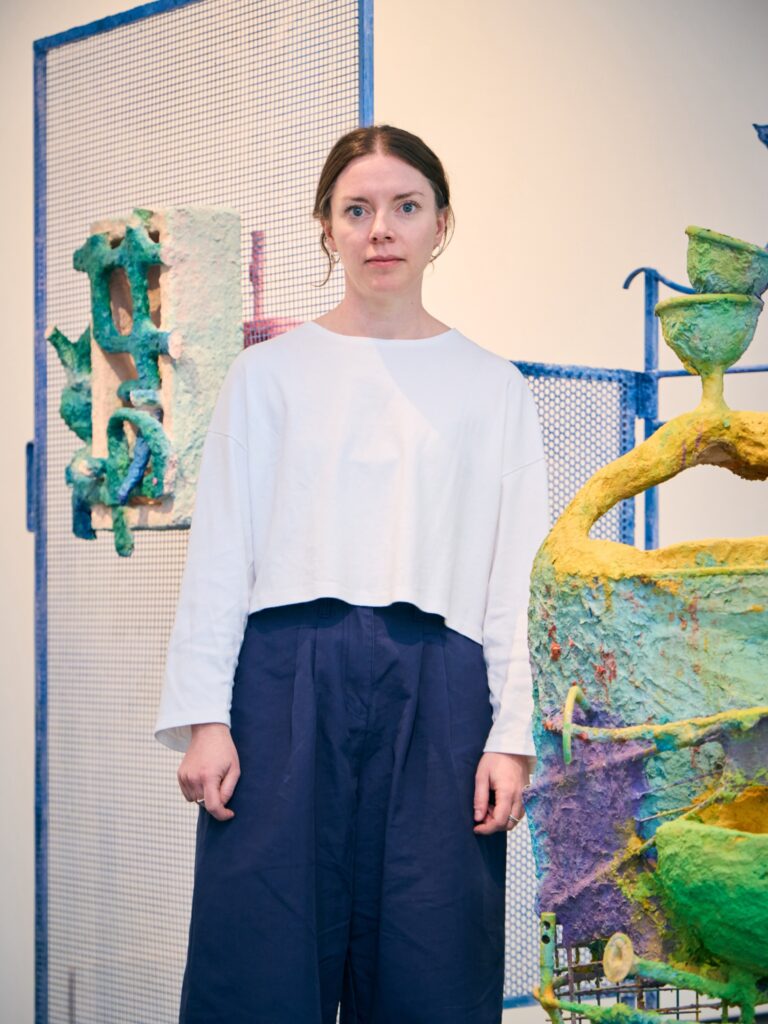 A woman with light brown hair tied back, wearing a white long-sleeve top and blue pants, stands in an art gallery with colorful abstract sculptures and mesh panels behind her.