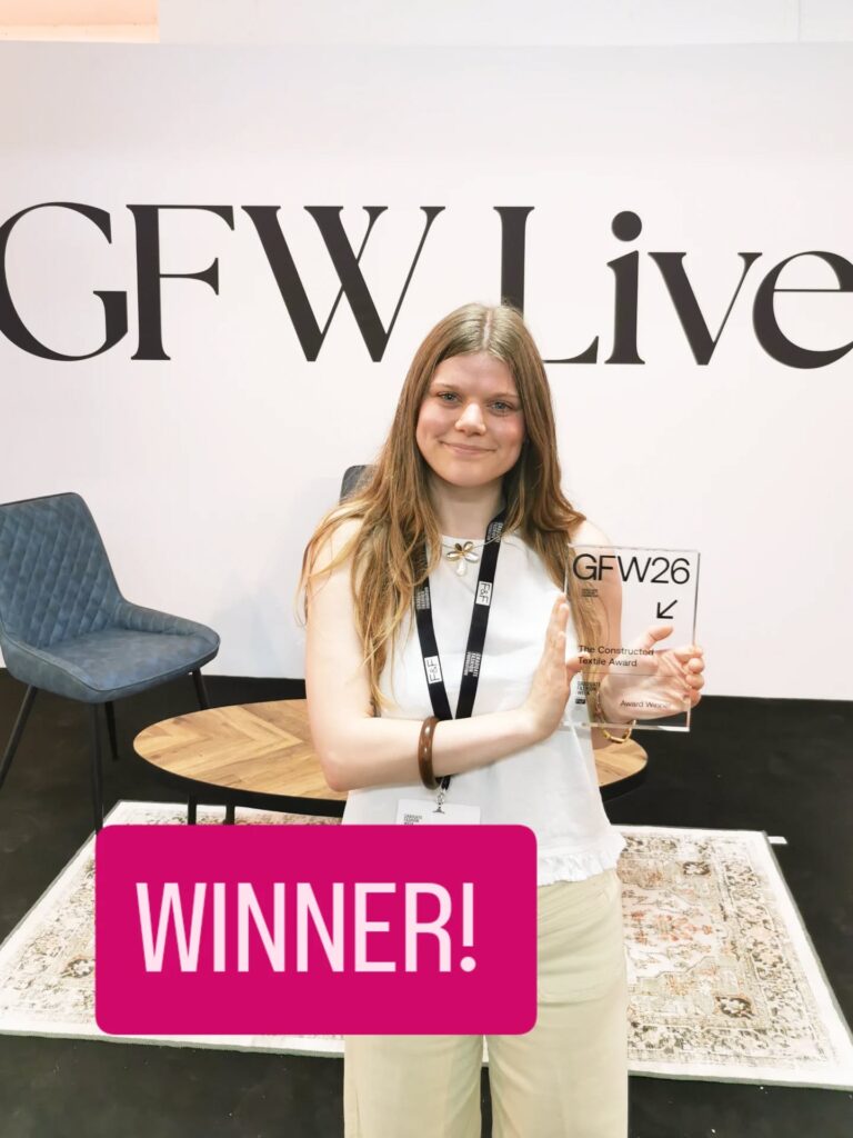 A smiling young woman holding a clear award plaque stands in front of a "GFW Live" sign. She wears a white sleeveless top and beige pants. The word "WINNER!" is displayed in bold pink text at the bottom of the image.