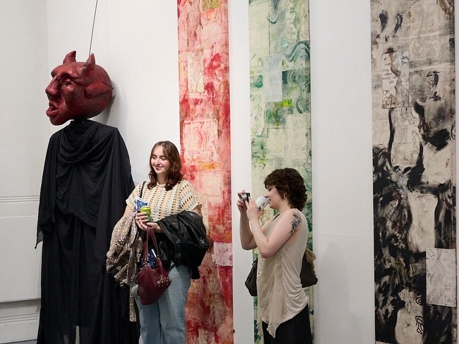 Two women stand in an art gallery; one smiles holding coats, the other takes a photo. Behind them are three vertical abstract artworks and a large red devil mask with a black cloak displayed on the wall.