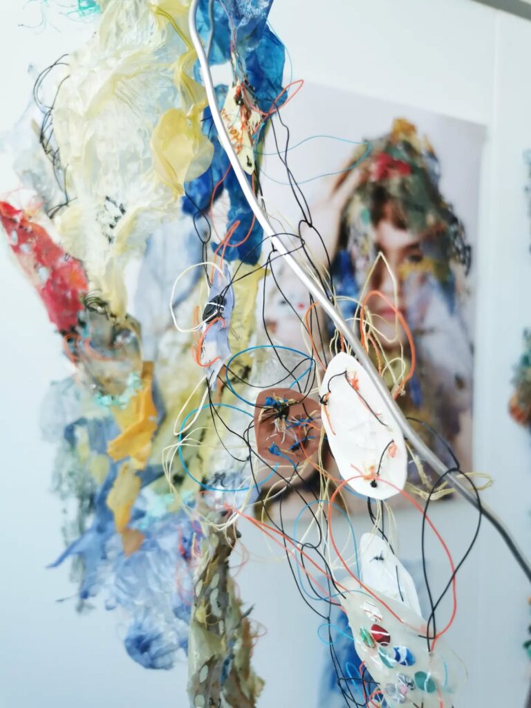 Close-up of colorful, tangled threads and translucent materials hanging, with a blurred image of a person in the background. The artwork has an abstract, textured appearance.