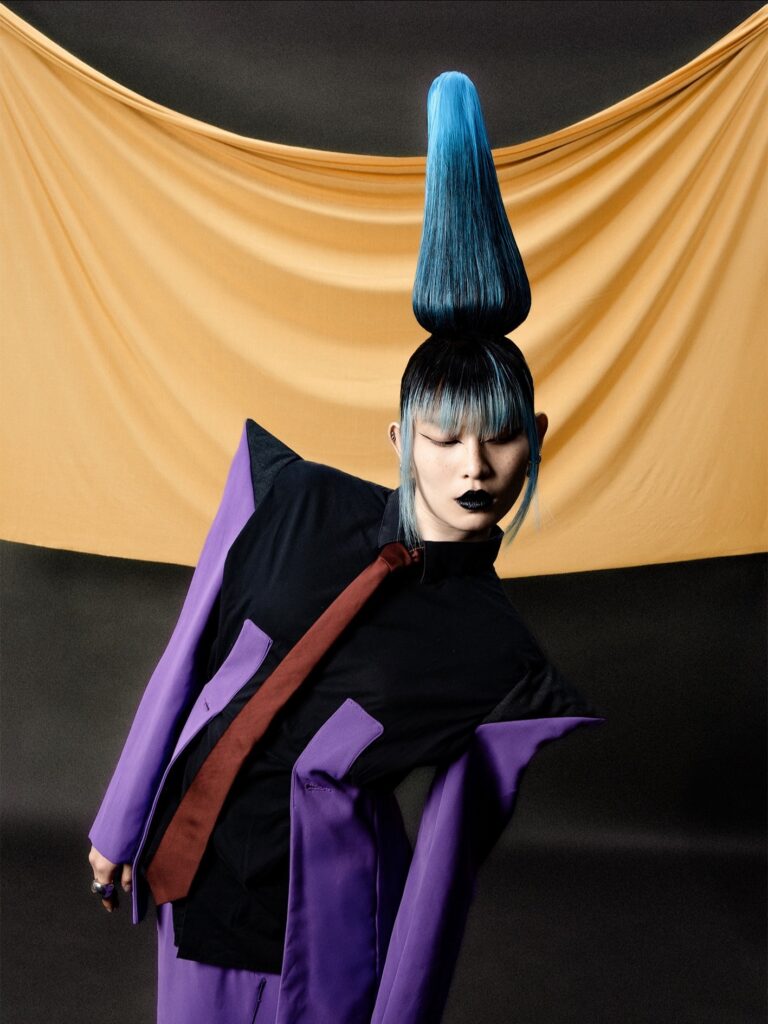 A person with dramatic, blue-tipped, cone-shaped hair wears a black shirt, red tie, and purple jacket with exaggerated shoulders, posed against a draped yellow cloth backdrop.