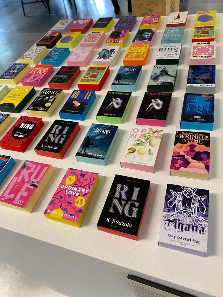 A display of colorful miniature books arranged in neat rows on a white table, showcasing various book covers and titles in different genres and styles.