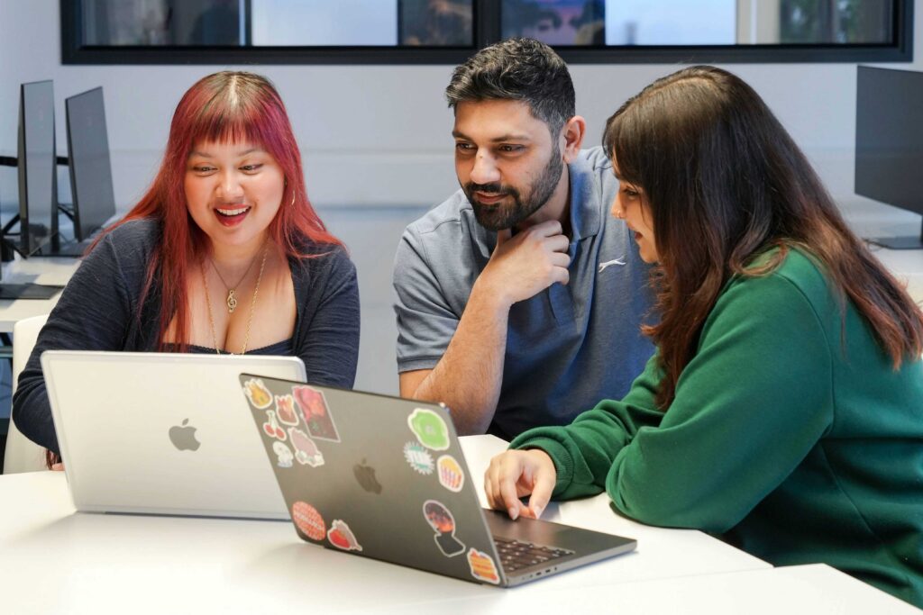 Three people sit at a table, smiling and looking at two laptops decorated with stickers in a modern, bright office or classroom setting with monitors in the background.
