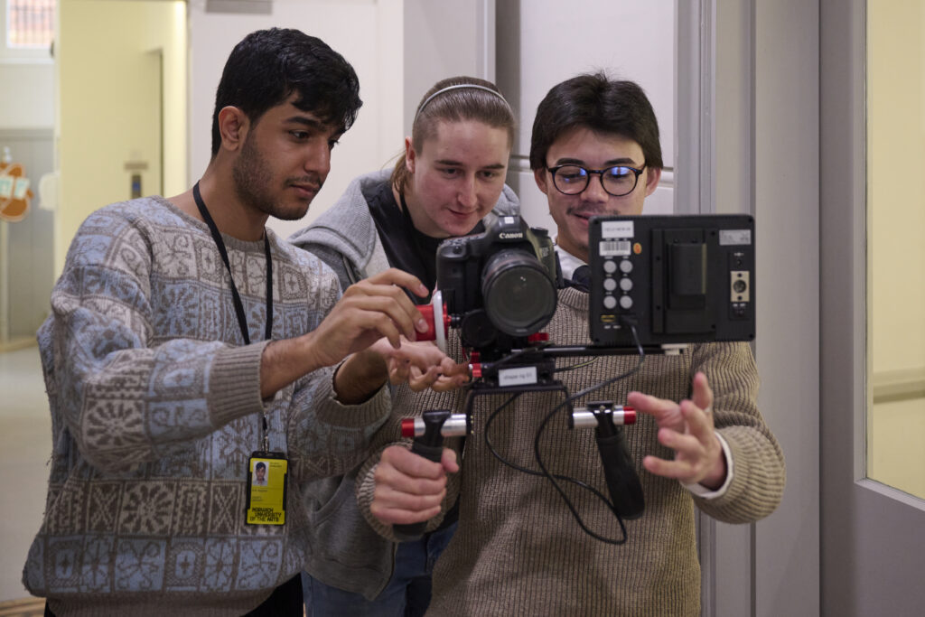 Three people stand indoors, focusing on a camera mounted on a stabilizer. One person holds the stabilizer while the others adjust the settings, collaborating closely and appearing engaged in a filmmaking activity.