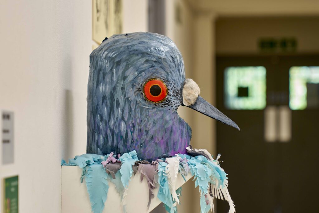 A large, colourful sculpture of a bird's head with vibrant orange eyes and a long beak sits on a shelf in a hallway, adorned with blue and purple fabric strips resembling feathers.