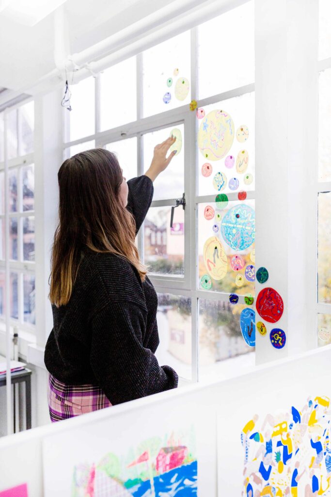 A person with long brown hair places a colorful circular sticker on a window decorated with various hand-drawn stickers in a bright, art-filled room. Artwork is also visible on the table in the foreground.