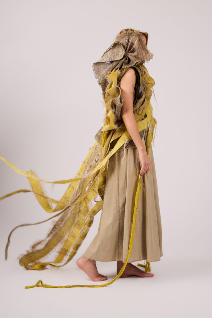 Person standing barefoot on a light background, wearing a loose beige skirt and a dramatic headpiece with layered, frayed yellow and brown fabric, some strips trailing behind them. The person is facing sideways.