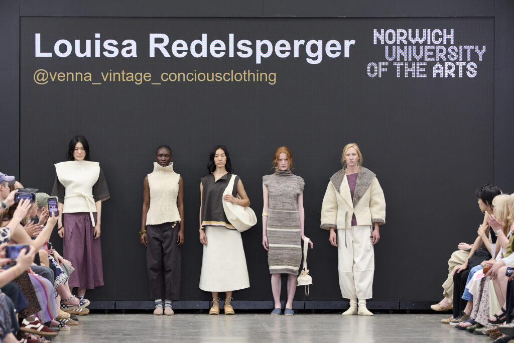 Five models stand in a line on a runway, wearing neutral-toned outfits. Behind them is a screen displaying "Louisa Redelsperger," social media handle, and "Norwich University of the Arts." An audience watches and takes photos.