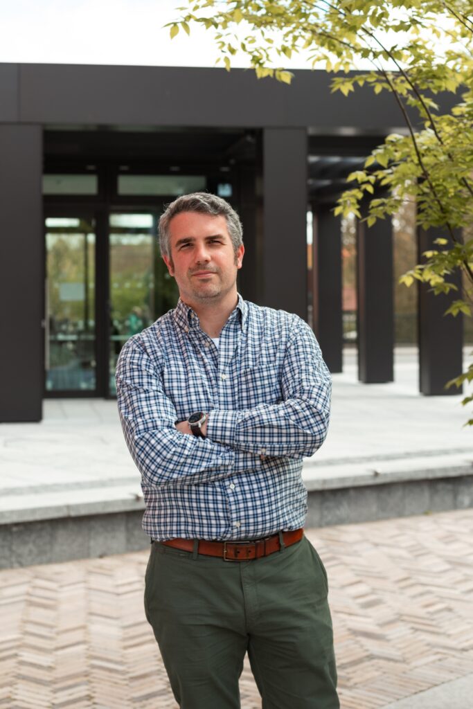A man with short dark hair, wearing a plaid shirt and green pants, stands outdoors with his arms crossed, in front of a modern building and leafy tree.