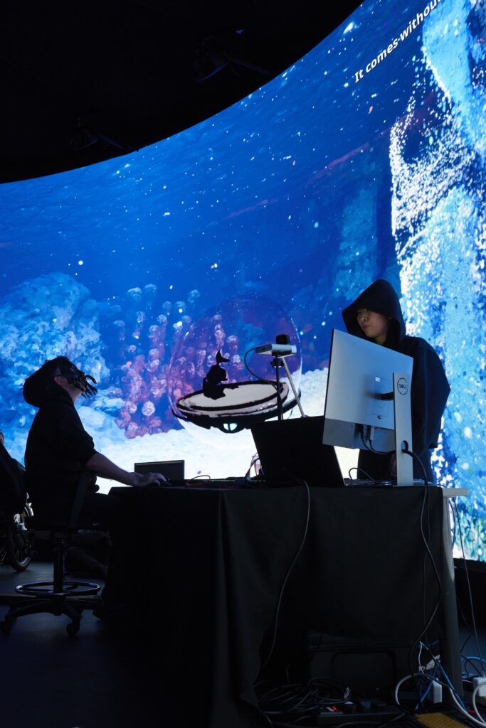 Two people work at computers in a dark room with a large, curved screen behind them displaying an underwater scene with coral and blue water. One person, wearing a hood, stands while the other sits facing the glowing underwater display.