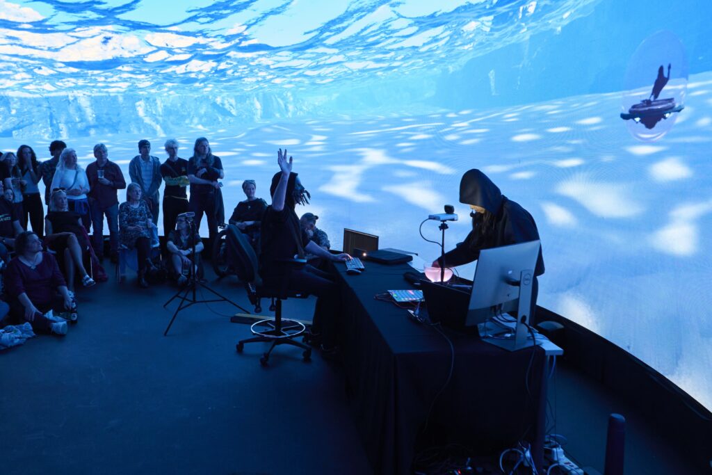 A person in a hoodie operates equipment at a table with computers while an audience watches. The room glows with blue underwater lighting effects, and a large curved screen displays an underwater ocean scene featuring a swimming seal.