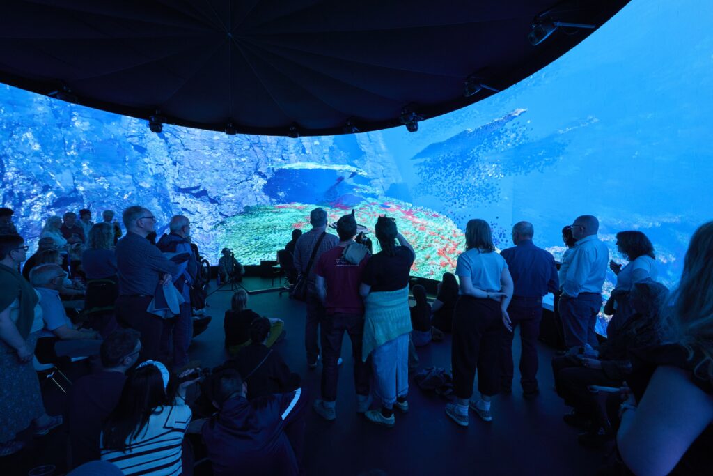 A group of people stand and sit inside a dark room, watching an immersive underwater aquarium display with blue water, fish, and colorful coral projected onto a curved screen.