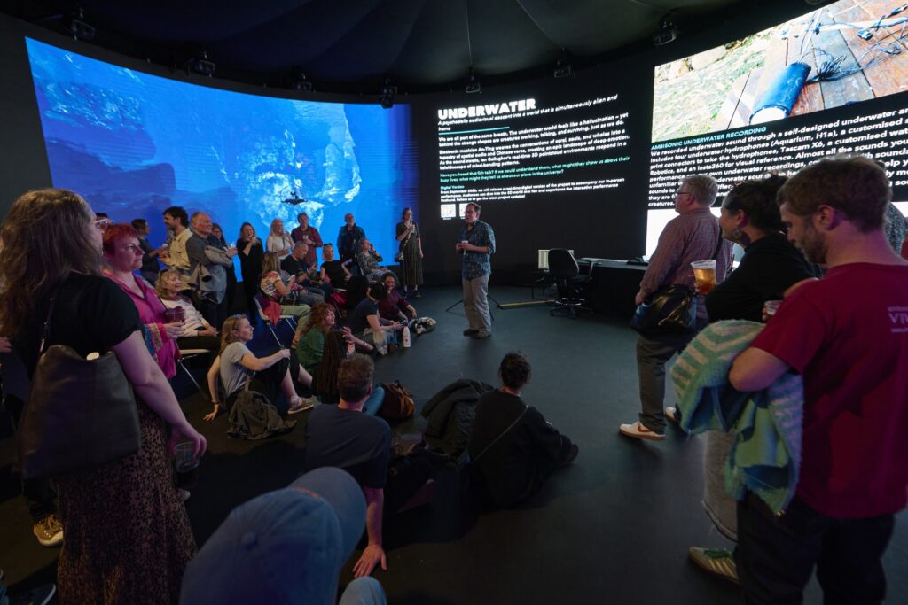 A group of people listen to a speaker in a dark room with large underwater-themed screens and text displays on the wall, creating an immersive underwater atmosphere. Some attendees are seated on the floor, while others stand around the presenter.