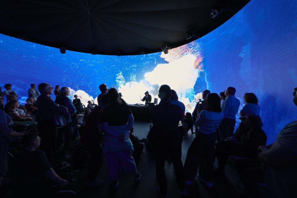 A crowd of people stands and sits in a dark room, facing a large curved screen displaying vivid underwater visuals in shades of blue, with a performer silhouetted on stage in front of the mesmerizing scene.