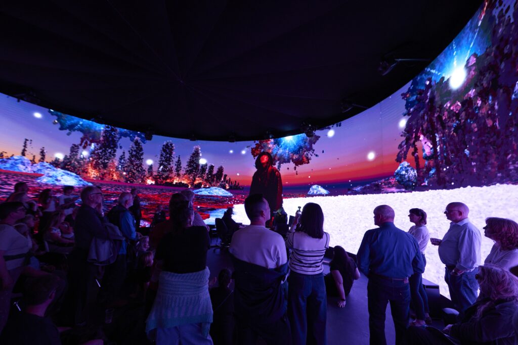 A group of people stand and sit inside a dark, circular room surrounded by immersive digital screens showing a vibrant, surreal landscape with underwater trees, snow, and a person in the center.