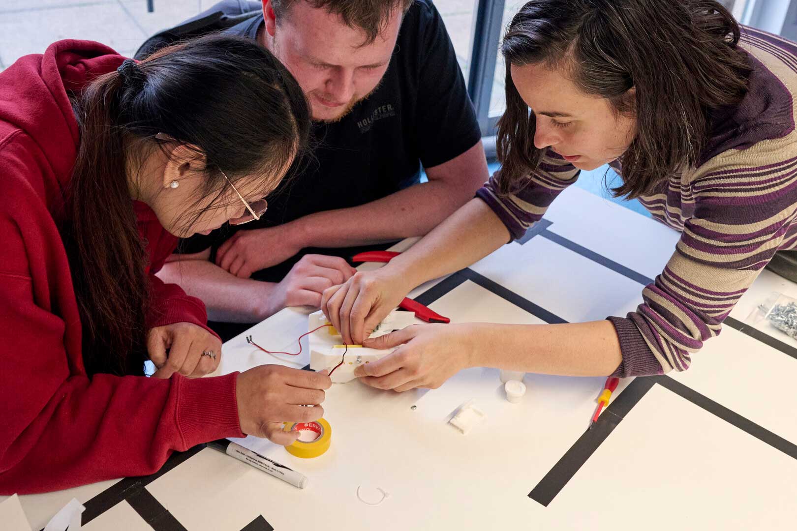 Three people work together on an electronics project at a table, handling wires and small components, with tools and materials scattered around them.