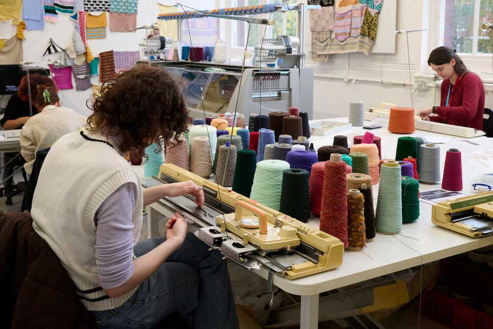 People sit at tables using knitting machines surrounded by colourful spools of yarn. Knitted samples hang on the walls in a bright, creative workspace.