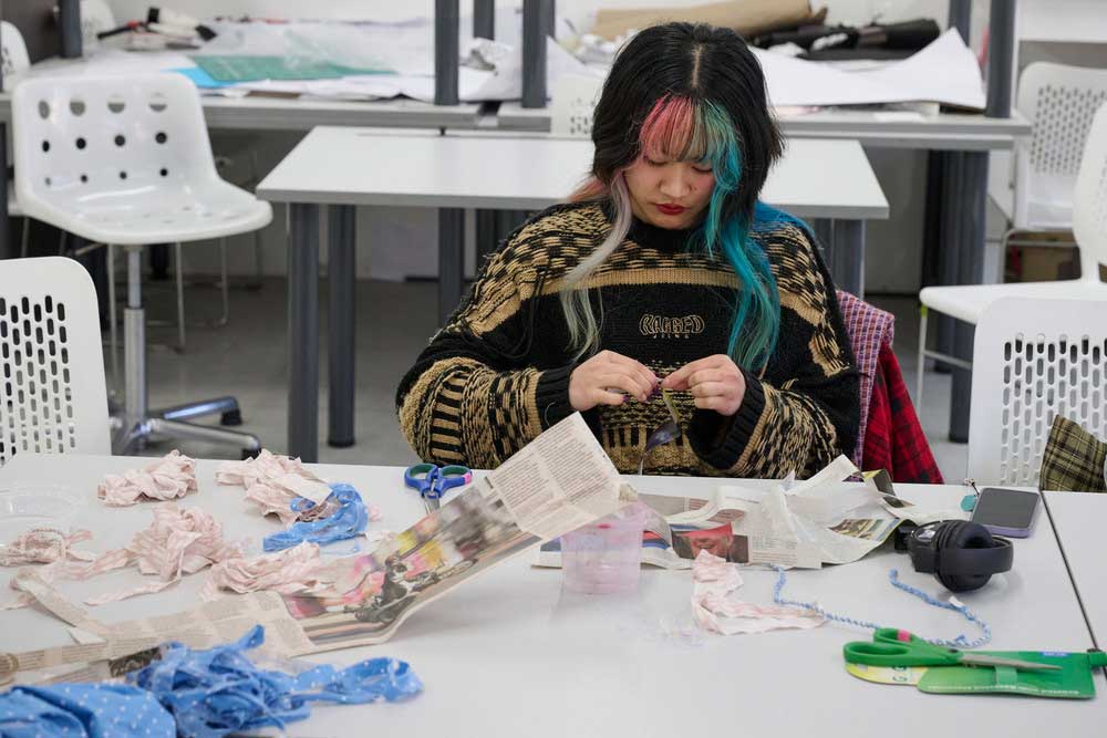 A person with multicoloured hair sits at a table, working with fabric strips and newspaper scraps. Scissors and other crafting materials are scattered around on the white table.