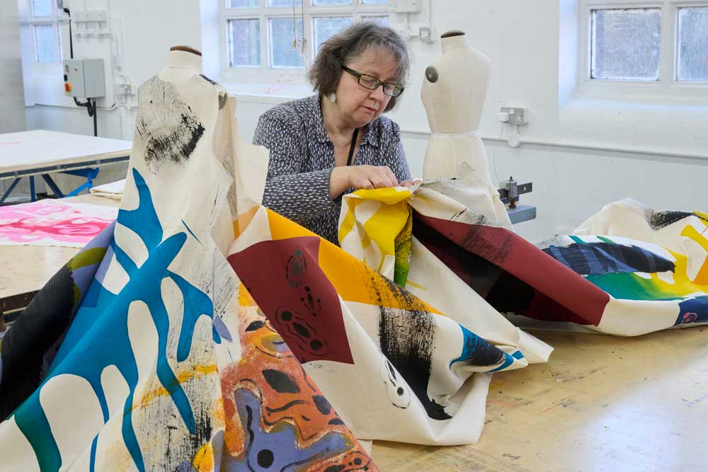 A person wearing a patterned shirt works with large pieces of colourful, patterned fabric at a table in a bright studio. Natural light streams in through the windows.