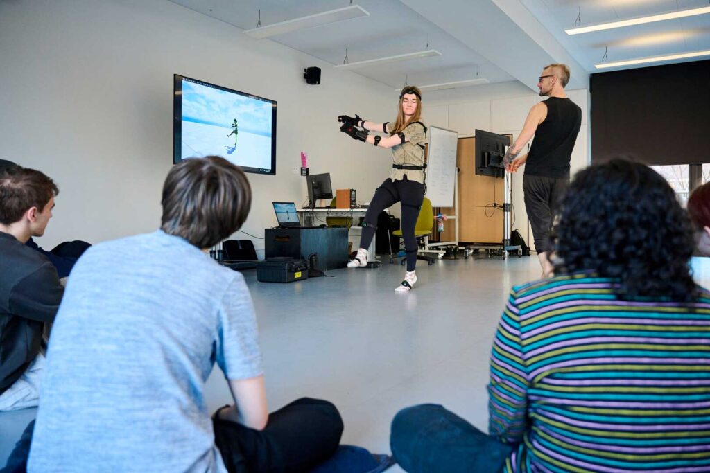 A person wearing motion capture gear demonstrates a pose in front of a screen showing an animated figure. A group of seated people observe the activity in a modern classroom.