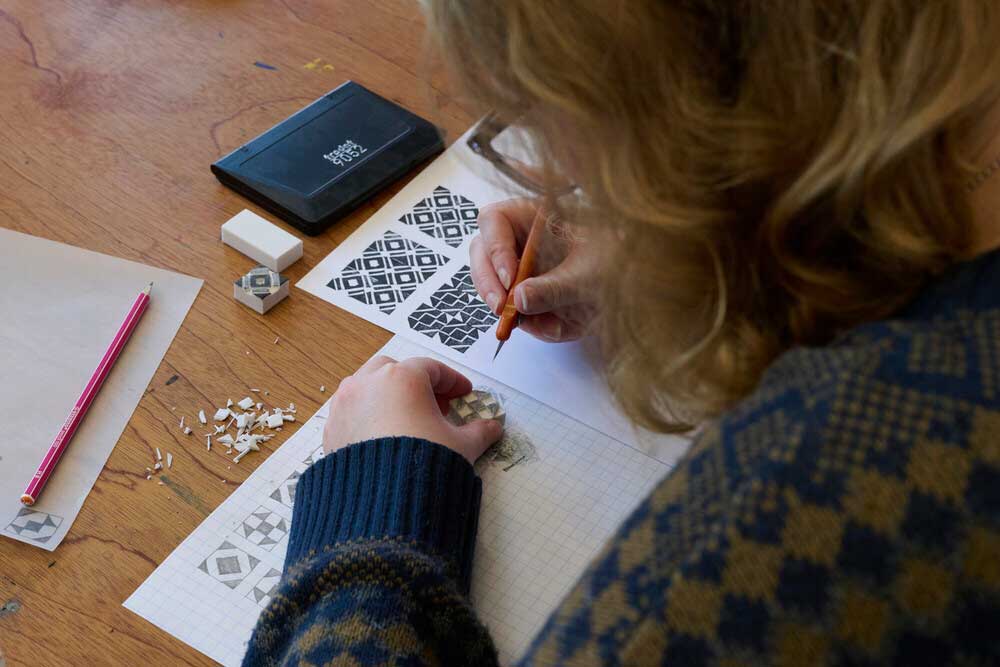 A person with blonde hair sketches geometric patterns on grid paper at a wooden table, surrounded by carving tools, patterned stamps and a pink pencil.