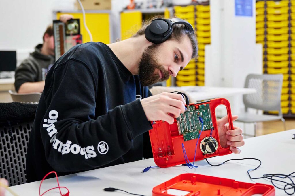 A person wearing headphones works on the circuit board inside a red plastic case, in a classroom or workshop setting.