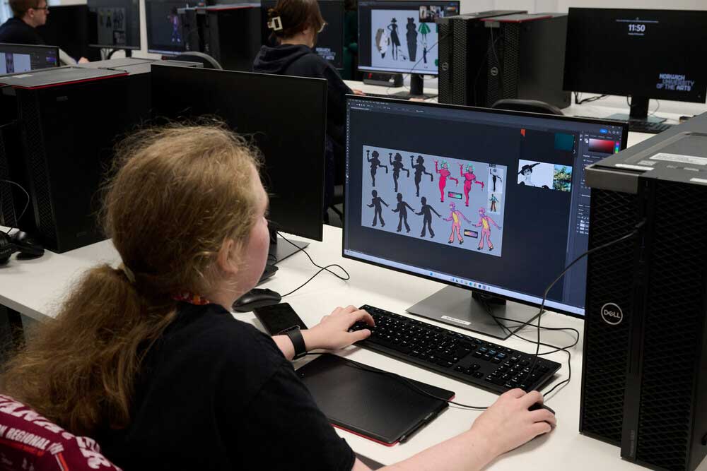 A person with long hair works at a computer in a lab, creating character designs on a screen. Other people are seated at computers in the background.