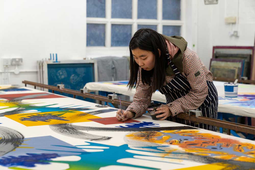 A person wearing a striped apron paints colourful, abstract shapes on a large canvas in a bright art studio.