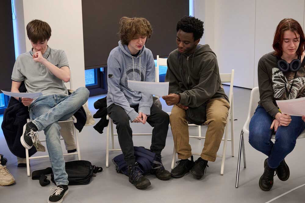 Four young people sit on chairs indoors, holding papers. The mood appears casual and focused.