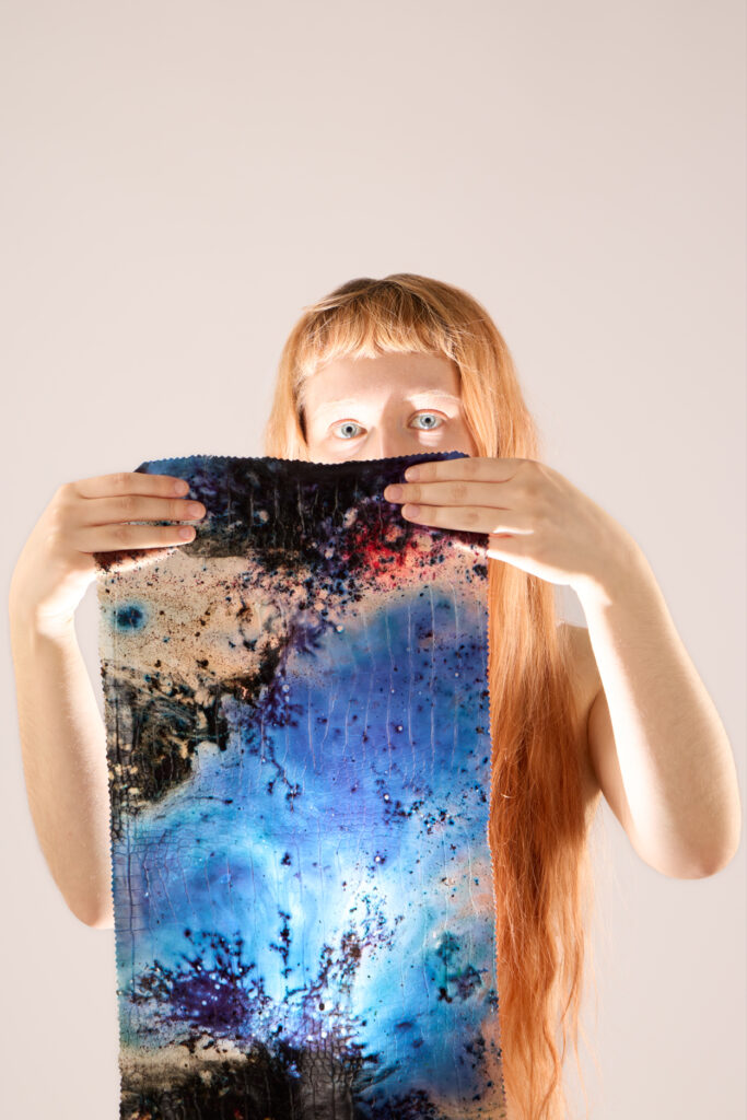 A person with long red hair holds up a colourful, abstract artwork featuring blue, black, and beige patterns, partially obscuring their face against a light background.