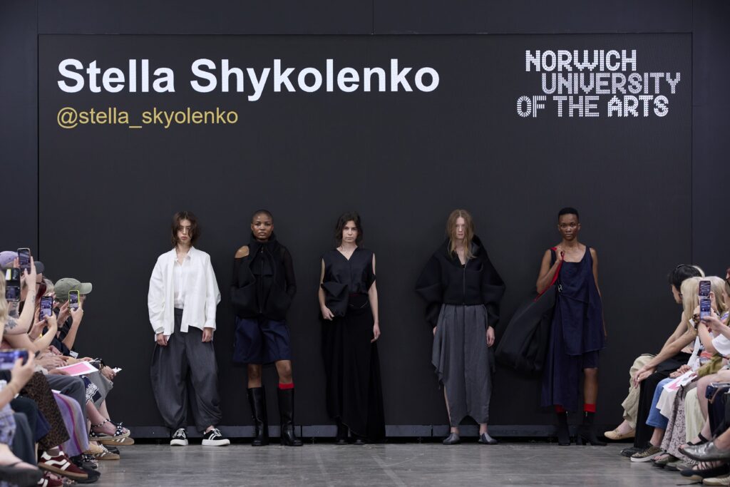 Five models stand on a runway wearing dark, modern outfits in front of a screen displaying "Stella Shykolenko" and "Norwich University of the Arts" as the audience takes photos.