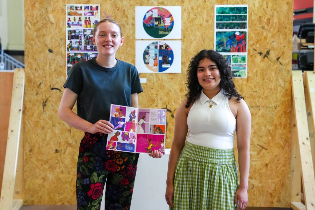Two young women stand indoors, one holding a colorful comic-style artwork. Behind them, a wooden display board shows more bright, graphic illustrations and art panels. Both are smiling and facing the camera.
