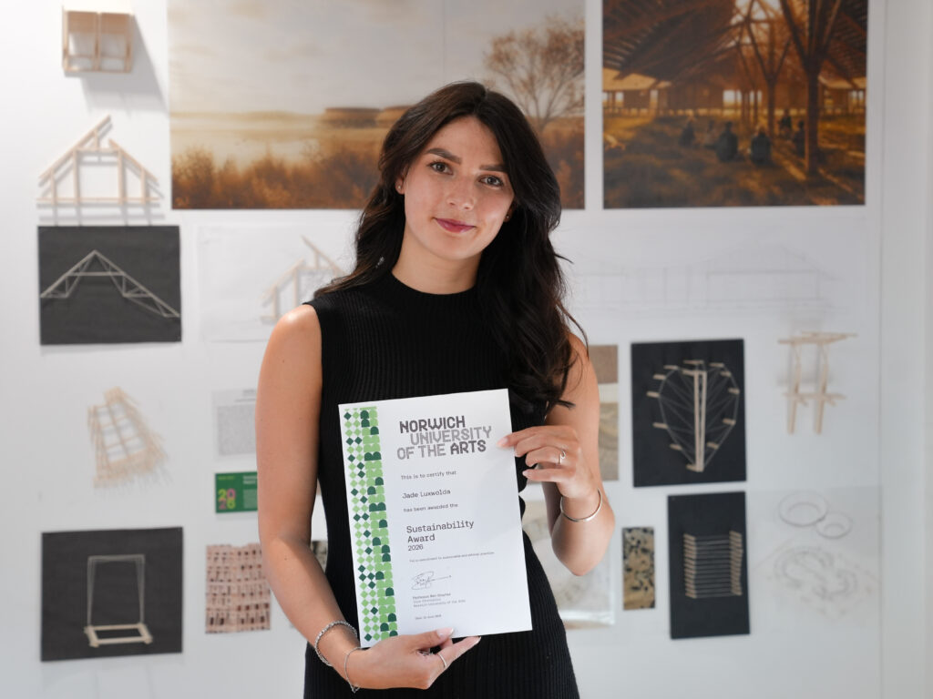 A person with long dark hair, wearing a sleeveless black dress, stands indoors holding a "Sustainability Award" certificate from Norwich University of the Arts. Architecture models and design posters are displayed behind her.