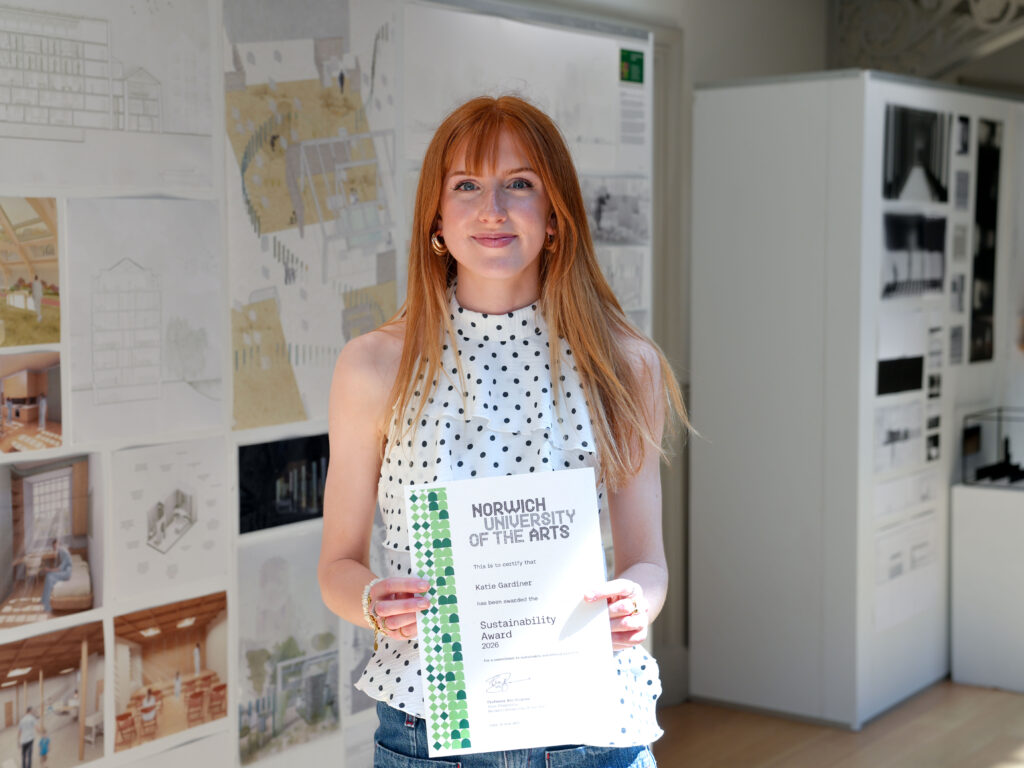 A person with long red hair, wearing a sleeveless polka dot blouse, stands indoors holding a Norwich University of the Arts certificate. Interior Design projects are displayed on the wall behind her.