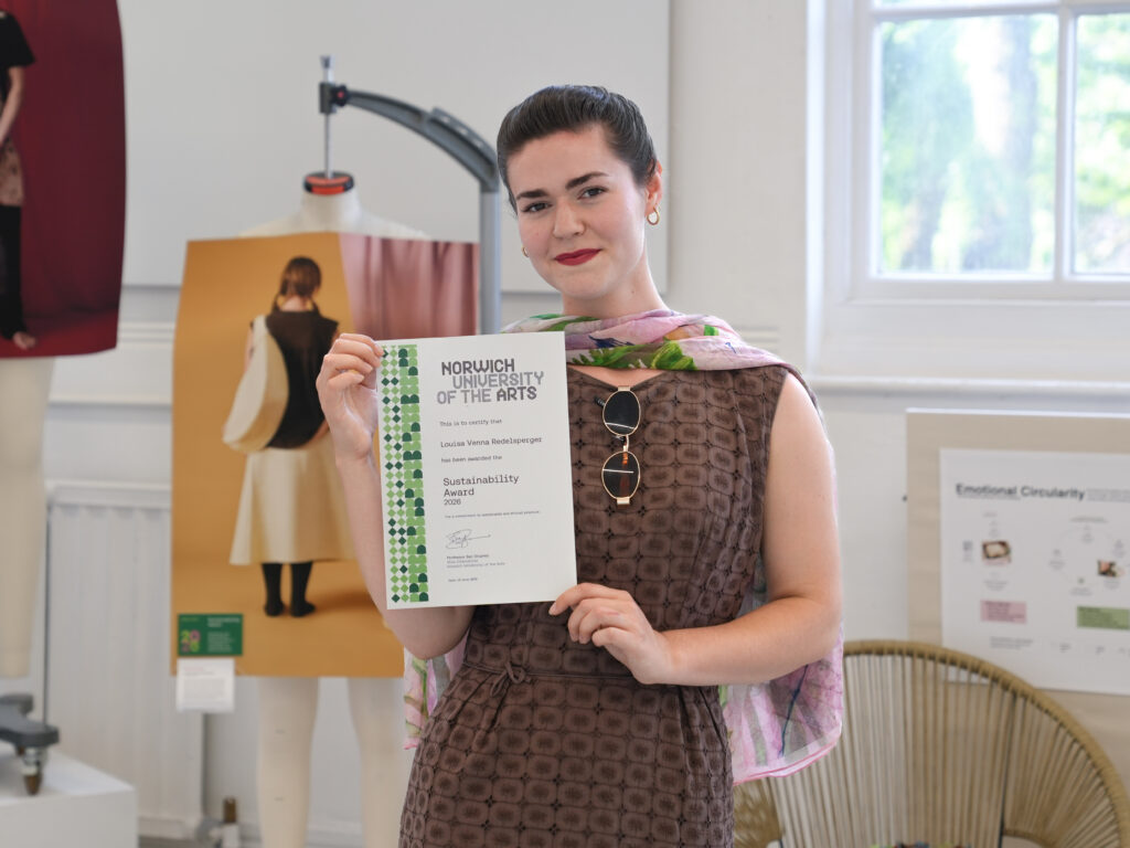 A person stands indoors holding a certificate from Norwich University of the Arts for a Sustainability Award. She is smiling and wearing a brown dress with a pink scarf, with art displays and a window in the background.