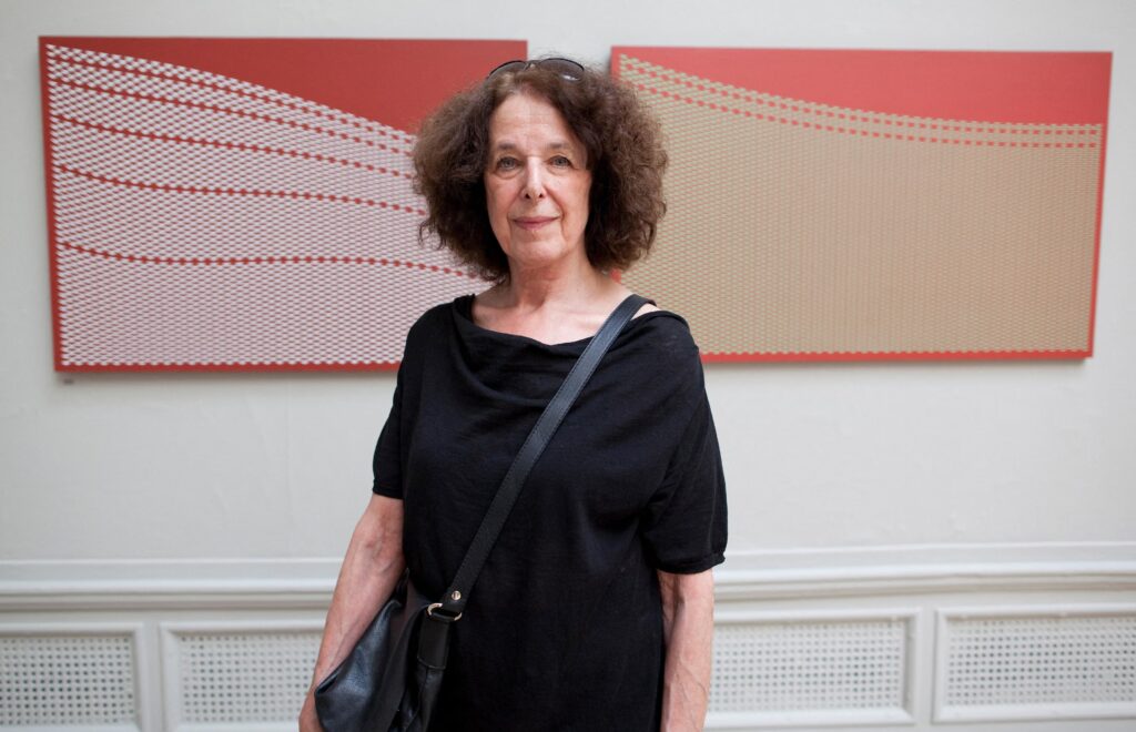 A woman with curly dark hair wearing a black top and shoulder bag stands in front of two abstract patterned artworks with red and beige tones mounted on a white wall.